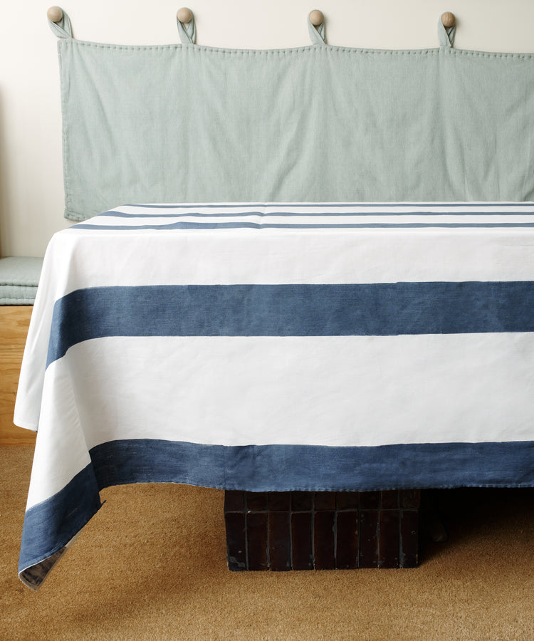 A table covered with the indigo-striped Riga Tablecloth sits on a brown carpet, with a light green cushioned bench against the wall in the background.