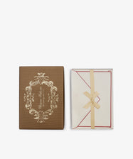 A set of 25 Bordered Correspondence Cards and Envelopes features a brown box adorned with an ornate gold design, placed next to an open cream envelope tied with a ribbon and showcasing red-bordered cotton paper inside—ideal for elegant wedding thank-you notes.