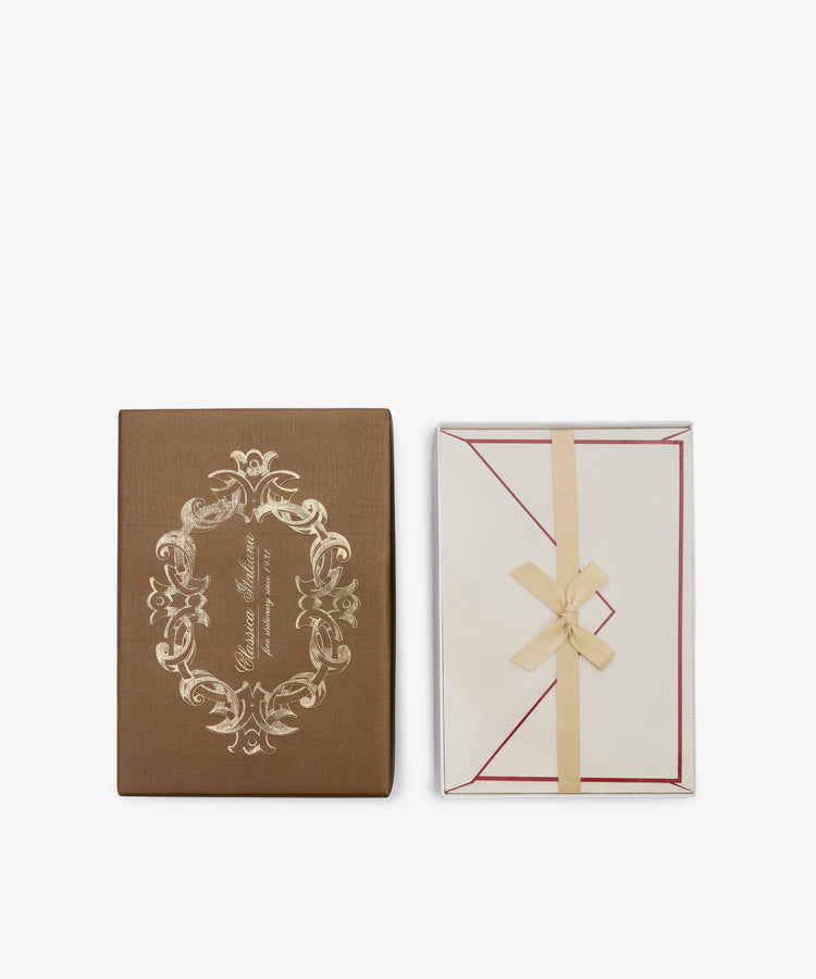 A set of 25 Bordered Correspondence Cards and Envelopes features a brown box adorned with an ornate gold design, placed next to an open cream envelope tied with a ribbon and showcasing red-bordered cotton paper inside—ideal for elegant wedding thank-you notes.