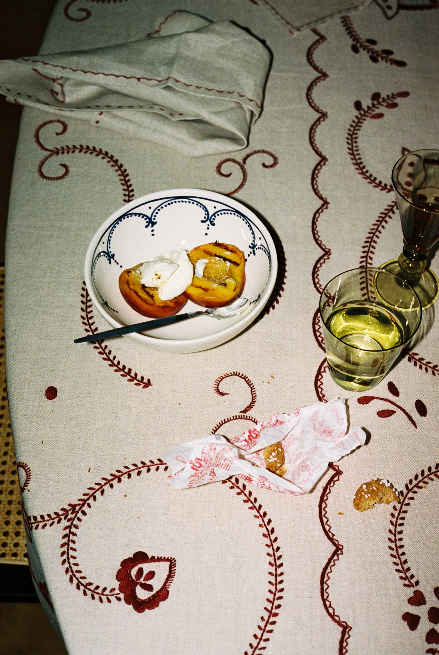 An Arco Cereal Bowl, handcrafted in Portugal, holds two peach halves and cream beside a glass of white wine, a wrapped cookie, and a glass of water on a patterned tablecloth.