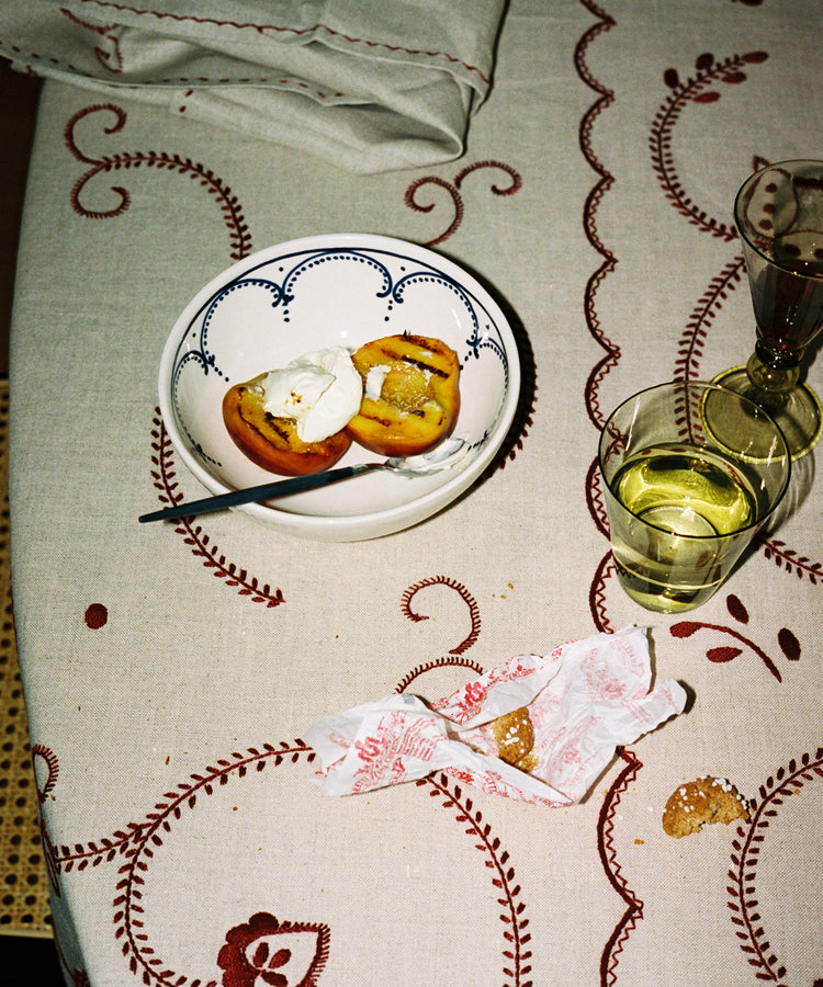 An Arco Cereal Bowl, handcrafted in Portugal, holds two peach halves and cream beside a glass of white wine, a wrapped cookie, and a glass of water on a patterned tablecloth.