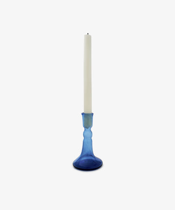 A white taper candle rests in the Lucia candleholder, made of cobalt glass, against a plain white background.