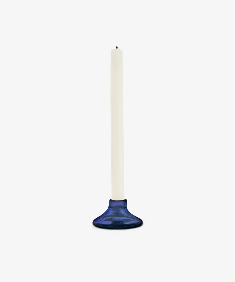 A white taper candle stands upright in the Lucien candleholder, Small—handblown in Morocco from recycled blue glass—against a plain white background.