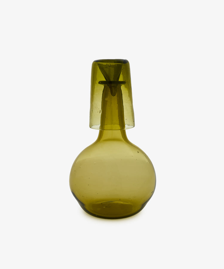 The Elodie Bedside Carafe and Glass Set features hand-blown green glass with a simple, rounded design and a slightly translucent finish; the matching cup inverts to rest atop the carafe, showcasing timeless elegance.