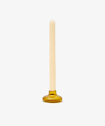 A single cream-colored taper candle sits in the Lucien candleholder, a round amber glass piece handblown in Morocco, set against a plain white background.