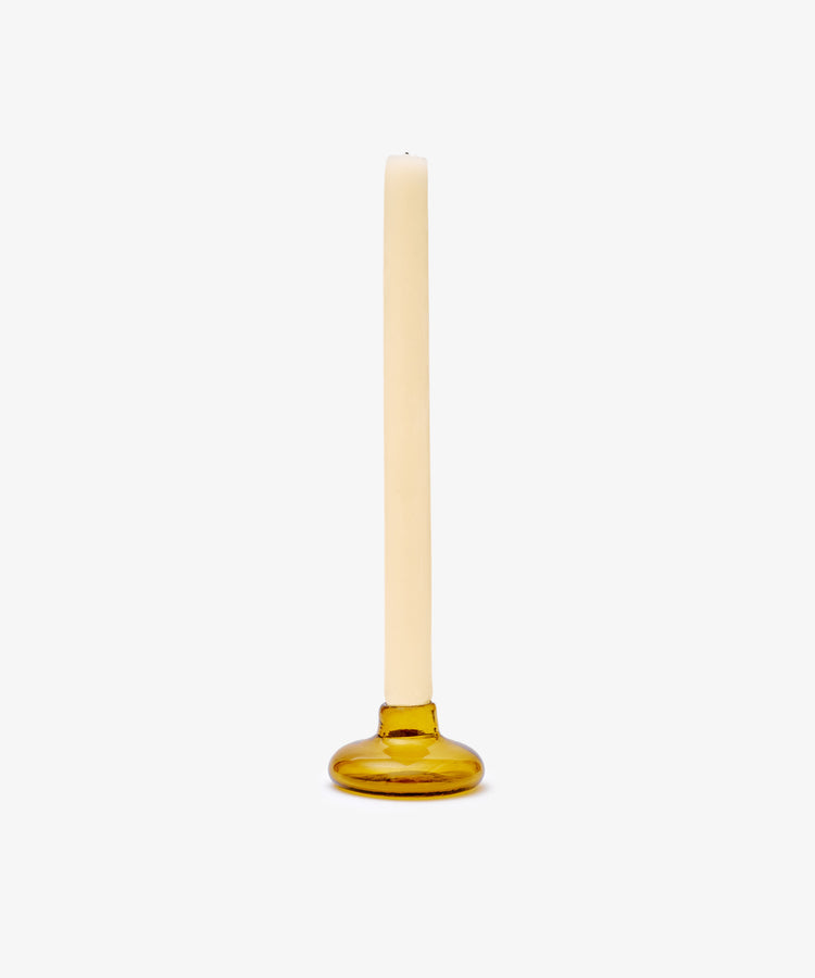 A single cream-colored taper candle sits in the Lucien candleholder, a round amber glass piece handblown in Morocco, set against a plain white background.