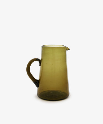 The Elodie Pitcher is a handblown, translucent olive-green glass pitcher with a handle and small spout, crafted in Damascus and shown against a plain white background.