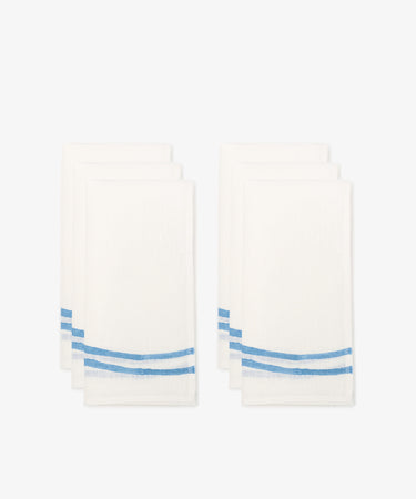Six Onda napkins, white linen with two blue wave-like stripes near one edge, arranged in two stacks against a plain white background.