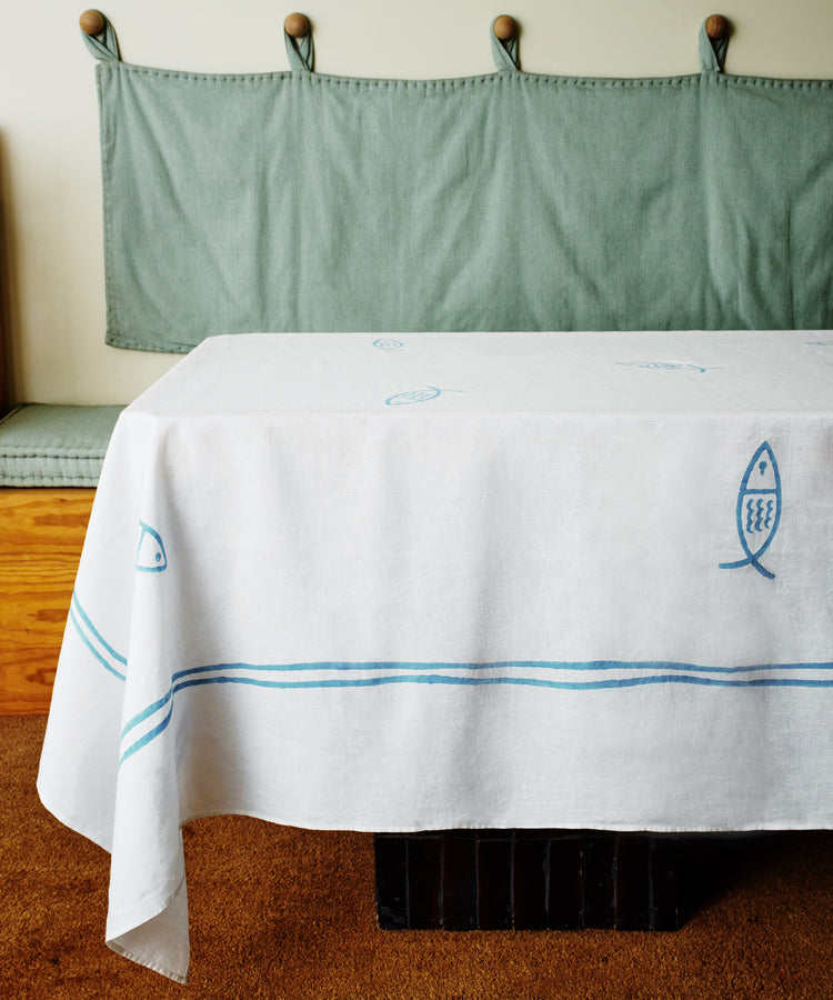 The Pezza Tablecloth, featuring handmade blue fish and line designs, covers a table in front of a bench with a green cushioned backrest.
