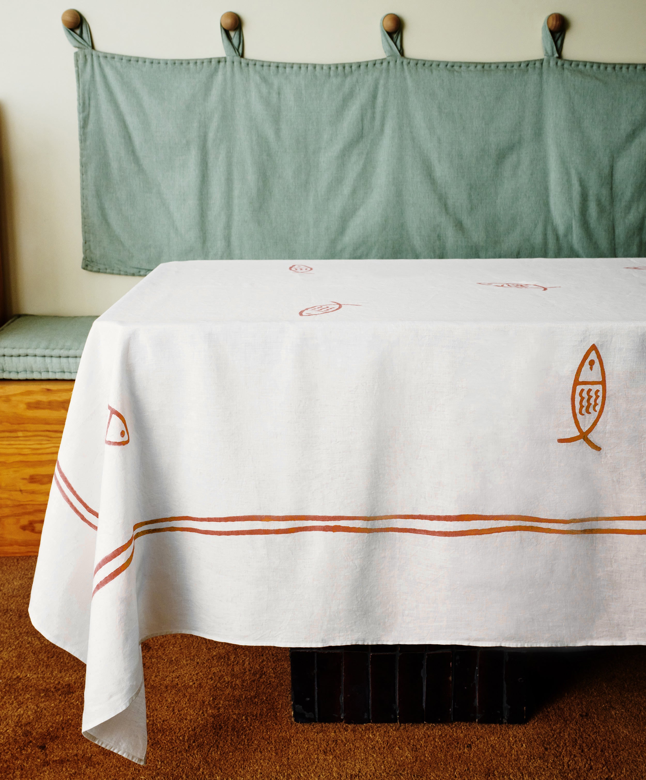 The Pezza Tableclothâ€”a white, hand block-printed tablecloth with red and brown fish and line designsâ€”drapes over a table beside a green cushioned bench with wall hooks, adding coastal charm to your dÃ©cor.
