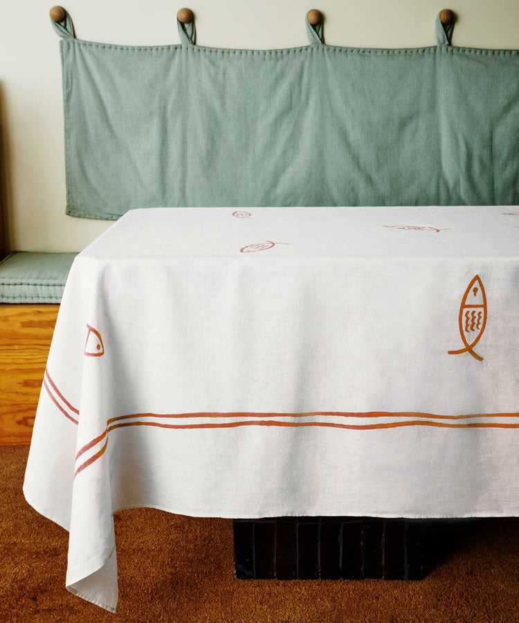 The Pezza Tableclothâ€”a white, hand block-printed tablecloth with red and brown fish and line designsâ€”drapes over a table beside a green cushioned bench with wall hooks, adding coastal charm to your dÃ©cor.