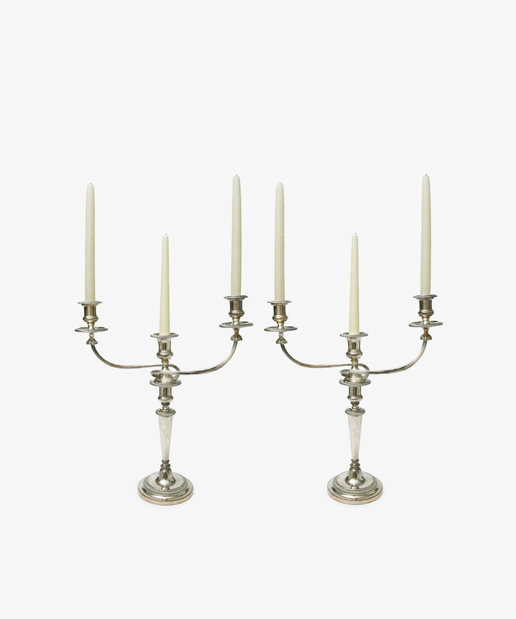 A set of two Vintage Silver-Plated Sheffield Plate Candelabra, each with three arms and holding tall unlit white taper candles, are shown against a plain white background, reflecting classic Georgian-era silversmithing.