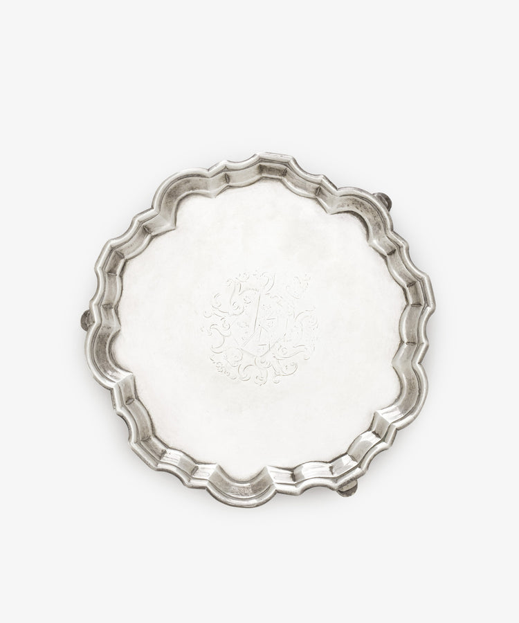 The Vintage English Salver is an 18th-century silver round tray with scalloped edges, three small feet, and a central engraved decorative pattern.