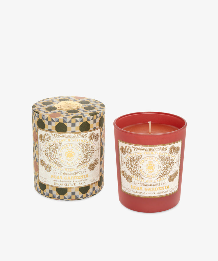 The Rosa Gardenia Candle, a red floral–musky candle in a glass holder with a decorative label, stands beside its patterned lid and container, evoking a velvety fragrance.