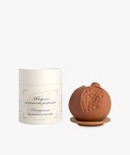 The Pomegranate in Scented Terracotta rests on a small dish beside a cream-colored Santa Maria Novella container with text and decorative accents, evoking the signature Melograno fragrance.