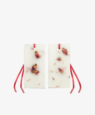 The Rosa Novella Scented Wax Tablets, Set of 2, are rectangular white wax tablets with embedded dried rose petals and red ribbon loops at the top.
