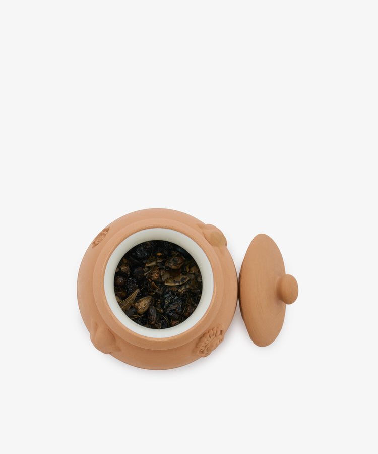 A small terracotta Pot Pourri jar with a white inner lining, filled with a traditional herbal blend, sits with its lid placed beside it on a white background.