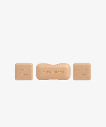 The Melograno Soap Set includes three beige bars from Santa Maria Novella on a white background: a rectangular center bar labeled “MELOGRANO” and two square side bars reading “SAPONE MELOGRANO.”.