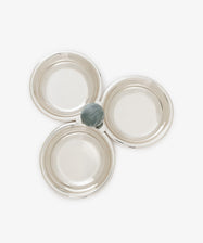 Three empty silver bowls from the Paros Triple Bowl Set are arranged in a triangular formation, with a single small round fruit—possibly a plum—placed at the center where the bowls converge, making it perfect for an elegant aperitivo presentation.