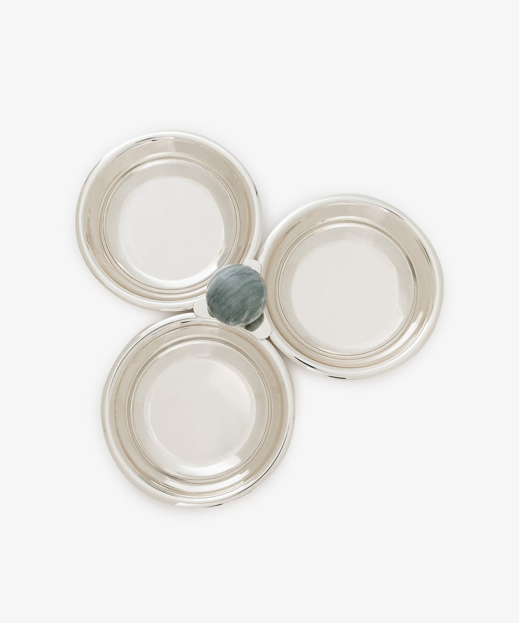 Three empty silver bowls from the Paros Triple Bowl Set are arranged in a triangular formation, with a single small round fruit—possibly a plum—placed at the center where the bowls converge, making it perfect for an elegant aperitivo presentation.