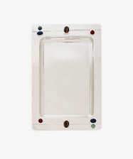 The Verola Rectangle Tray is a polished silver tray with rounded corners, adorned with four colored accents—two red, one green, and one blue stones or glass pieces—set near the edges. This decorative tray is presented on a white background.