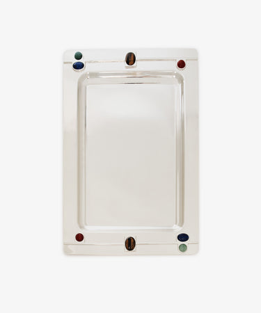 The Verola Rectangle Tray is a polished silver tray with rounded corners, adorned with four colored accents—two red, one green, and one blue stones or glass pieces—set near the edges. This decorative tray is presented on a white background.