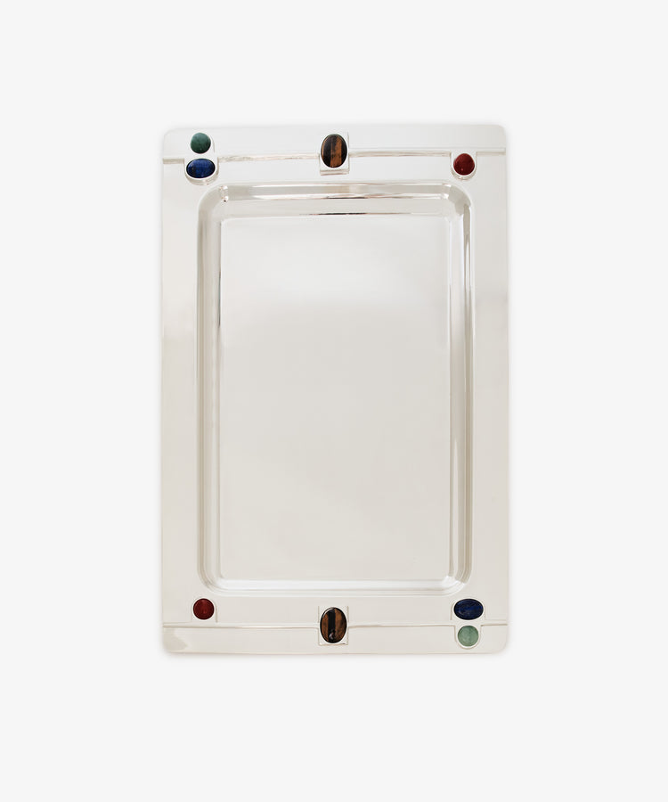 The Verola Rectangle Tray is a polished silver tray with rounded corners, adorned with four colored accents—two red, one green, and one blue stones or glass pieces—set near the edges. This decorative tray is presented on a white background.