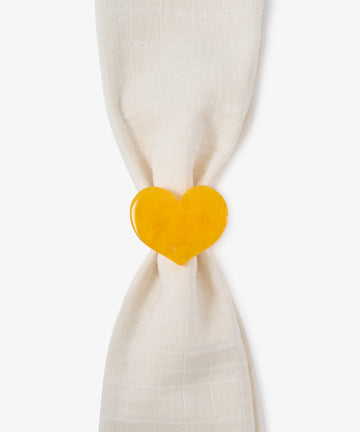 A cream-colored cloth napkin is gathered by the Heart Resin Napkin Ring, its sculptural, yellow heart form elegantly expressing love against a white background.