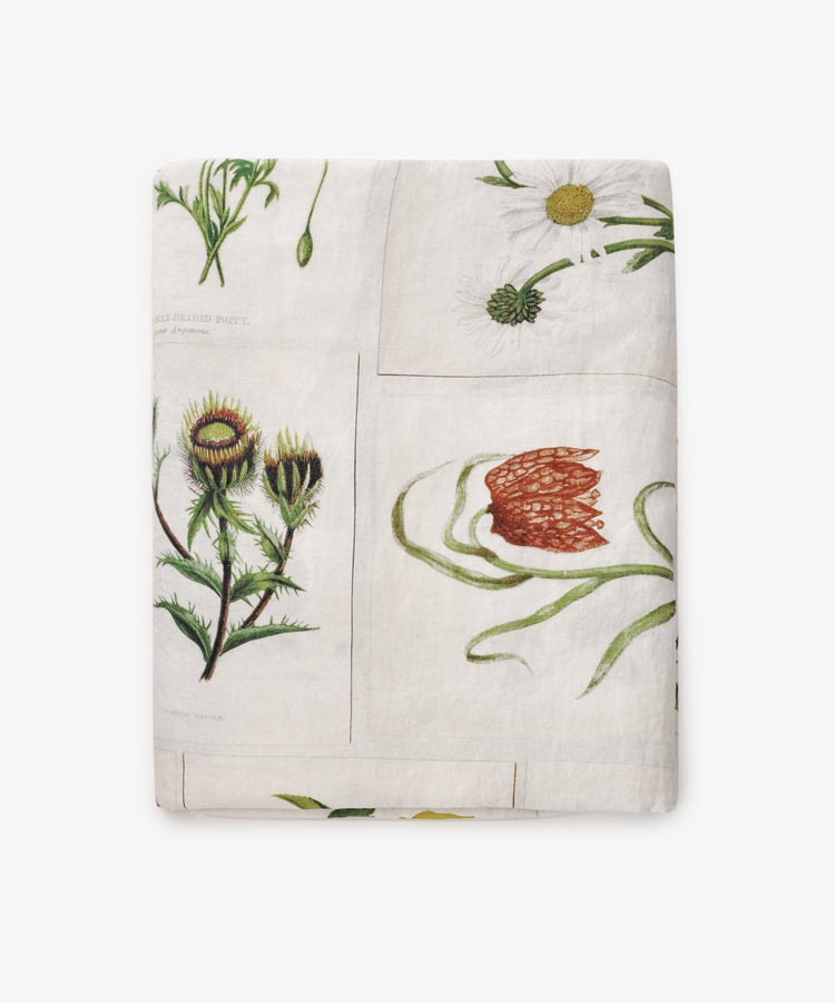 The John Derian Botanical Tiles Tablecloth is a folded linen tablecloth with botanical illustrations of red, white, and green flowers on a white background, showcasing John Derianâ€™s signature botanical design.