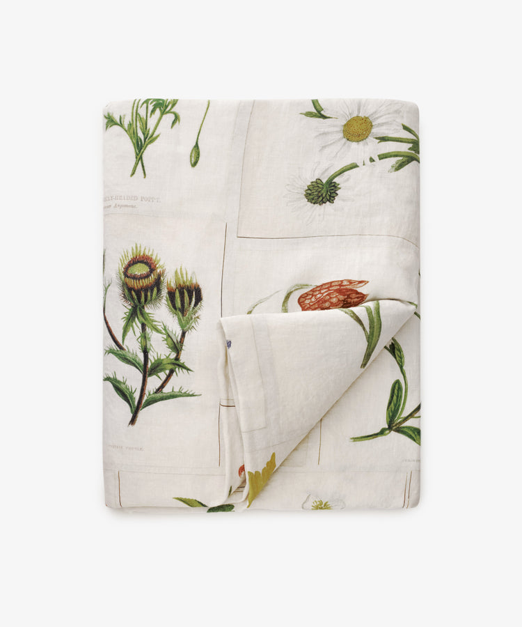 The John Derian Botanical Tiles Tablecloth is a cream-colored linen piece, folded and adorned with illustrated flowers and plants in a botanical design.