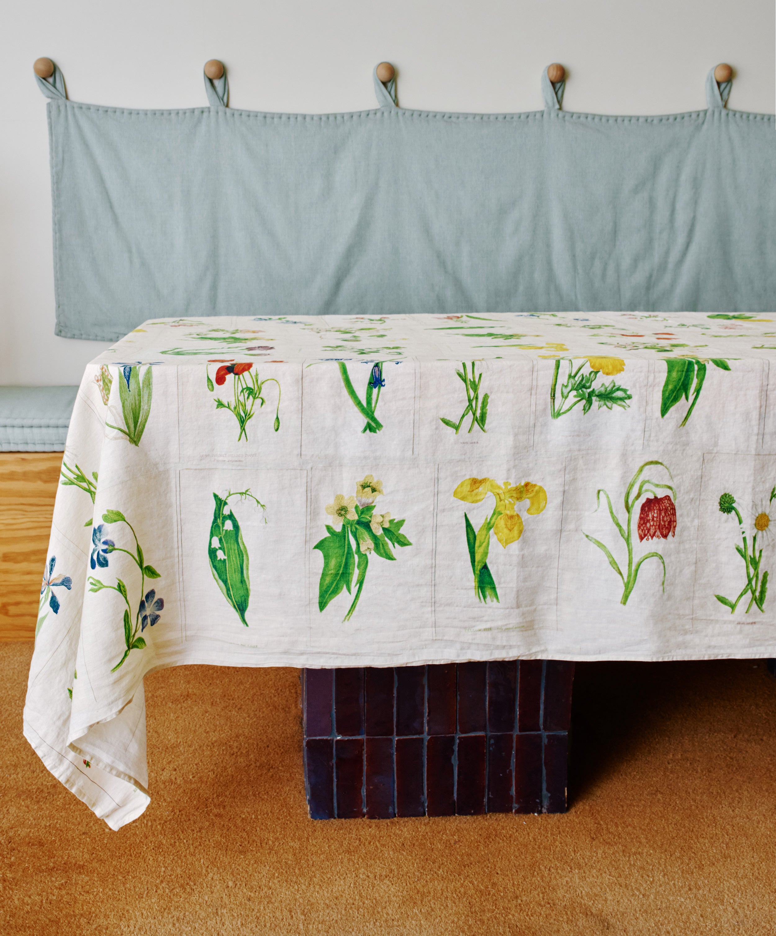 A table covered with a John Derian Botanical Tiles Tablecloth featuring botanical prints stands in front of a cushioned bench with a light blue fabric backrest.