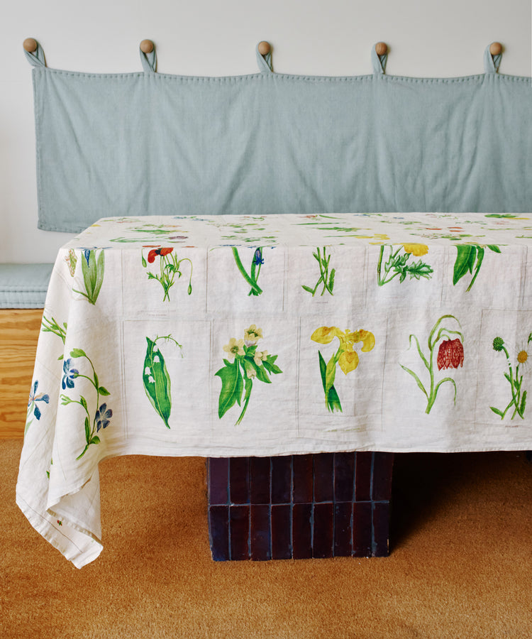 A table covered with a John Derian Botanical Tiles Tablecloth featuring botanical prints stands in front of a cushioned bench with a light blue fabric backrest.