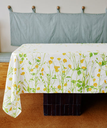 A Buttercup Linen Tablecloth with yellow flowers and green stems covers the table in front of a cushioned bench featuring a blue cover and hooks above.