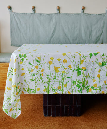 A Buttercup Linen Tablecloth with a yellow and green floral pattern covers the table, set before a light blue cushioned bench and beige wall hooks for charming garden-inspired decor.