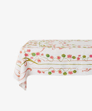 The Capucines Tablecloth, rectangular and white with a pink, green, and gold floral-vine Art Nouveau pattern, drapes elegantly over the table against a plain background.