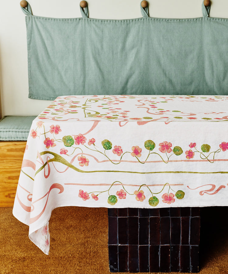 A table with the Capucines Tablecloth, featuring pink and green floral patterns, sits in front of a cushioned bench with a light green backrest.