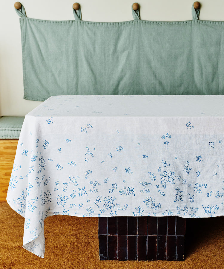 A table with the Falling Flower Linen Tablecloth, featuring blue hand-drawn flowers, stands in front of a bench with a green fabric backrest on a brown carpeted floor.