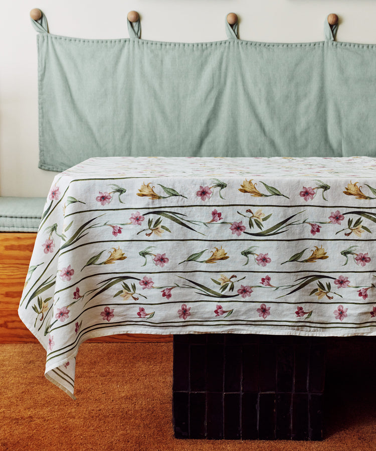 A table with a Gladioli Tablecloth sits in front of a bench featuring light green cushions and a wooden base, all atop a brown carpet.