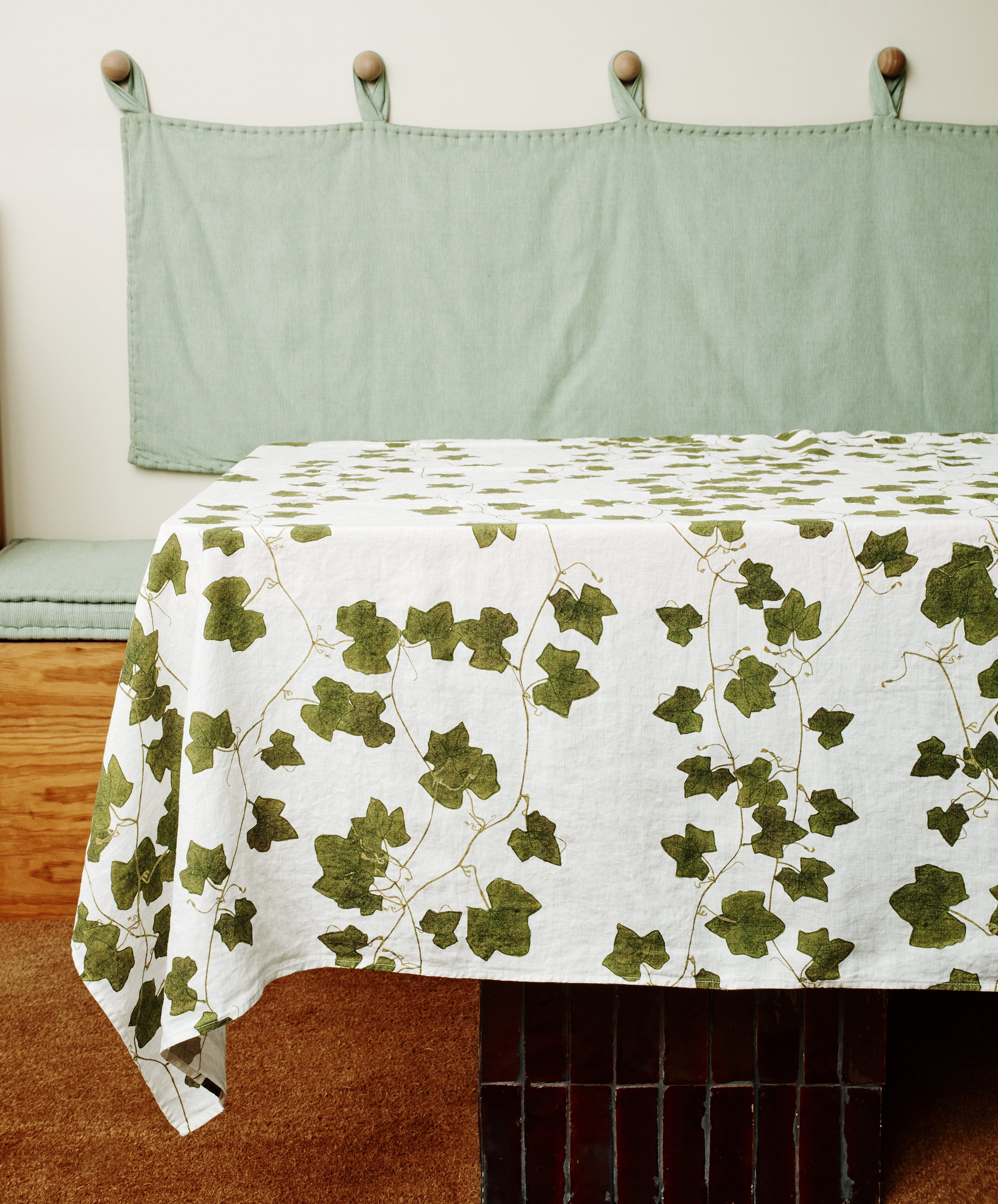 A table covered with the Ivy Herbarium Linen Tablecloth, Medium, and matching napkins is set in front of a green cushioned bench with a fabric backrest in a minimalist room.