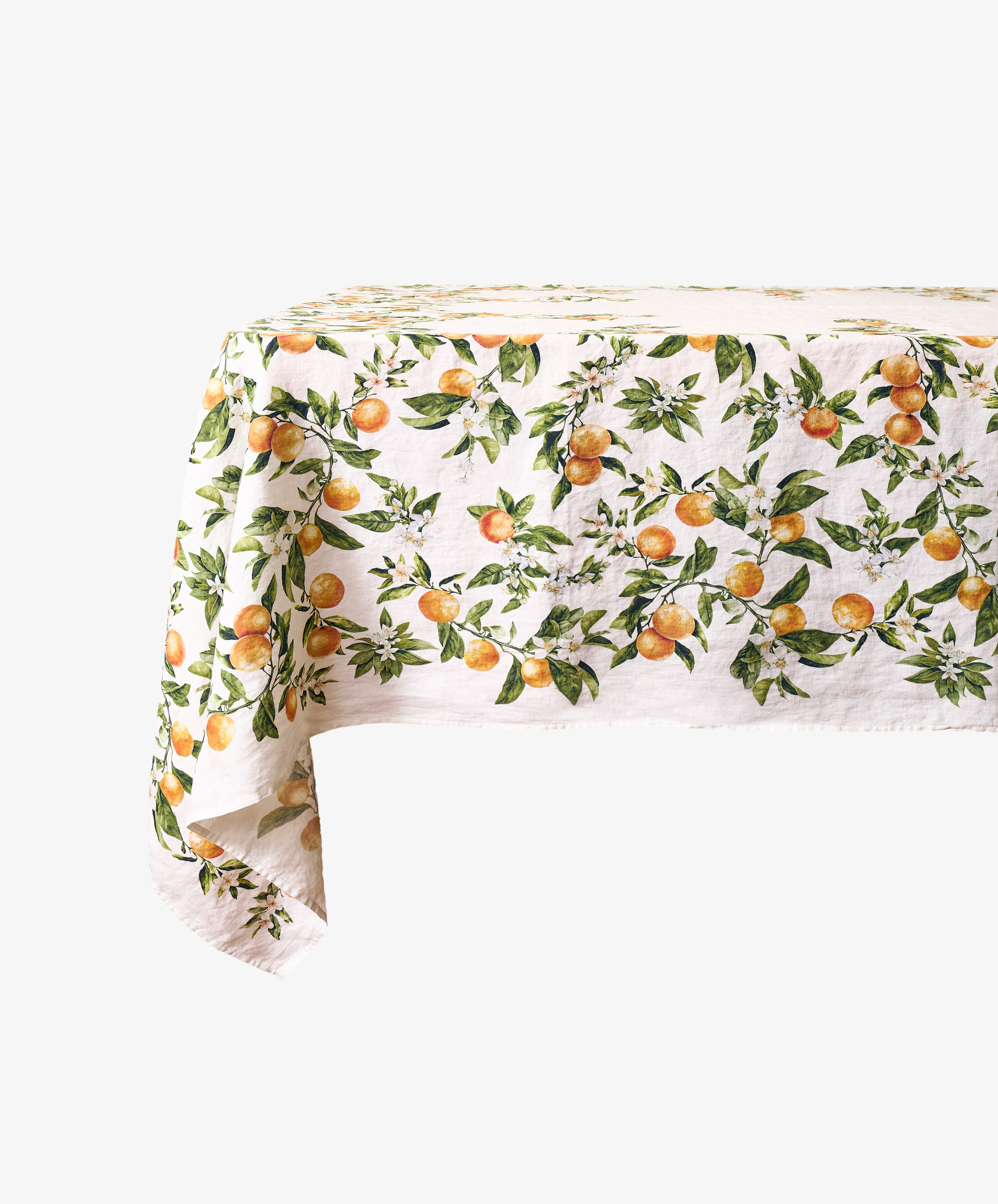 The L’Orangerie Linen Tablecloth, featuring an orange and green fruit pattern evocative of a classical French garden, is draped over the edge of a rectangular table set against a plain background.