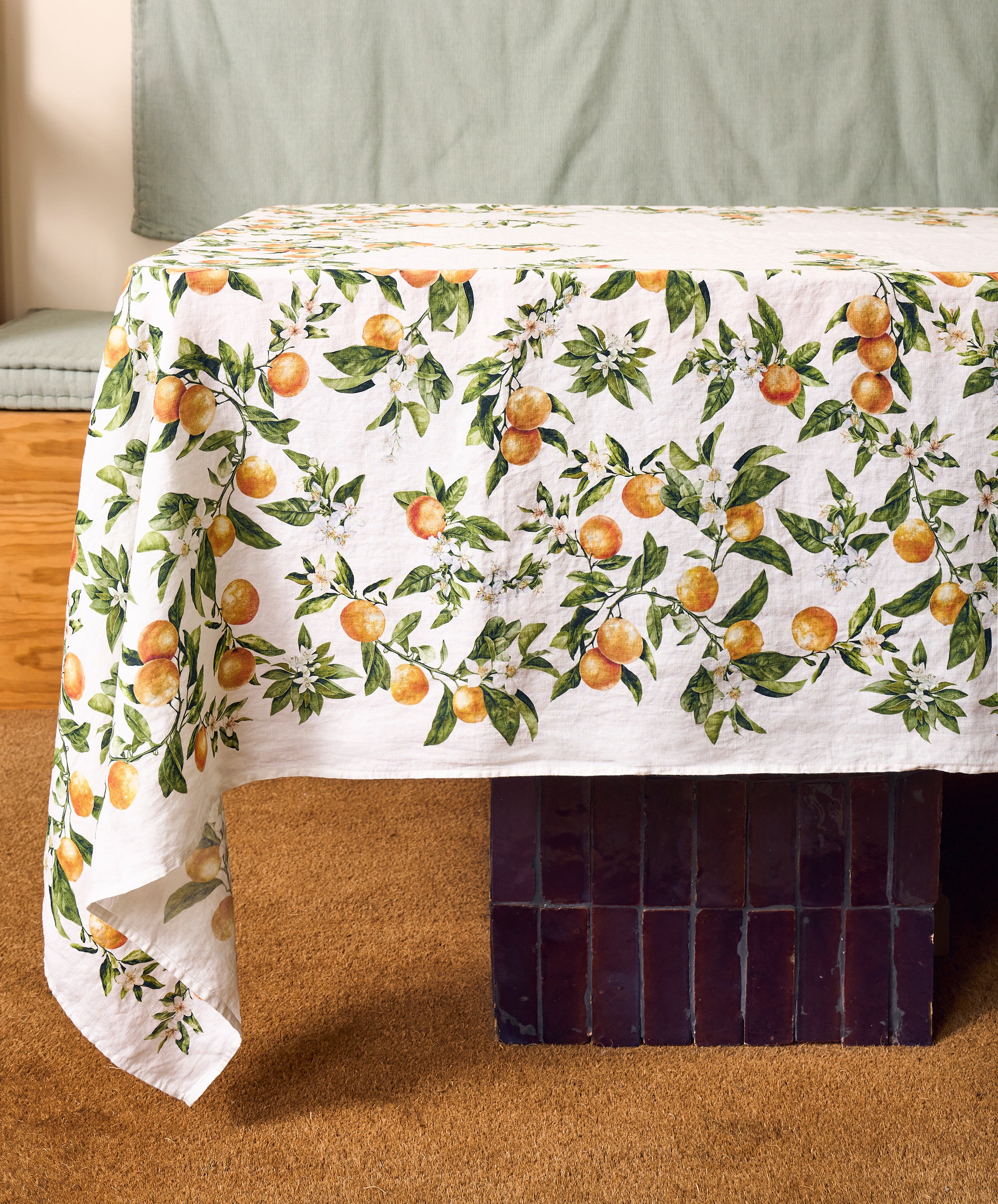 A rectangular table is covered with the L’Orangerie Linen Tablecloth—white French linen adorned with an orange fruit and green leaf pattern—and set in a room with brown carpet and a cushioned bench in the background, evoking the charm of a classical French garden table setting.