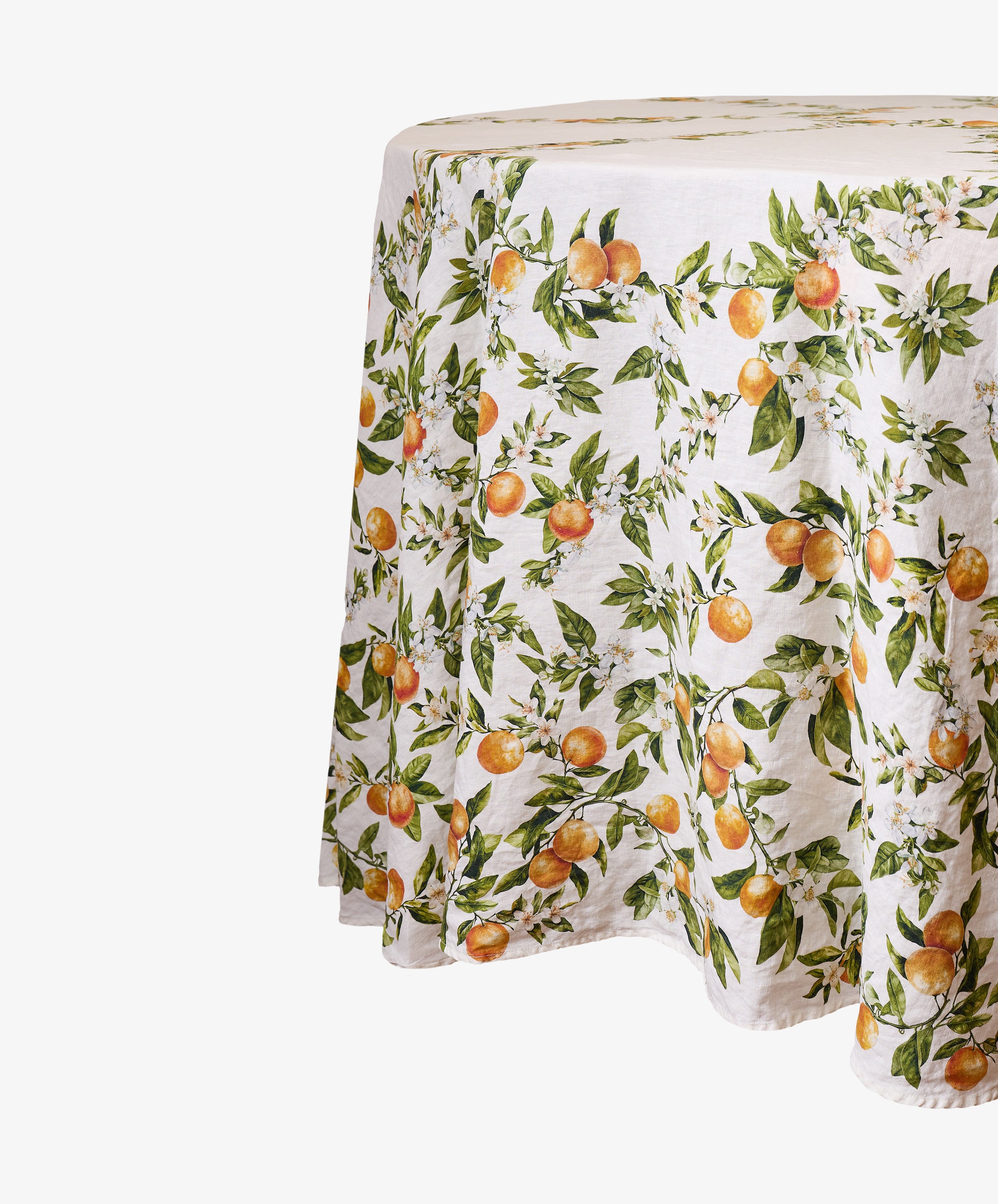 The L’Orangerie Round Linen Tablecloth covers the table with a white linen fabric adorned with a vibrant pattern of oranges and green leaves on branches, evoking classic French garden-inspired designs.