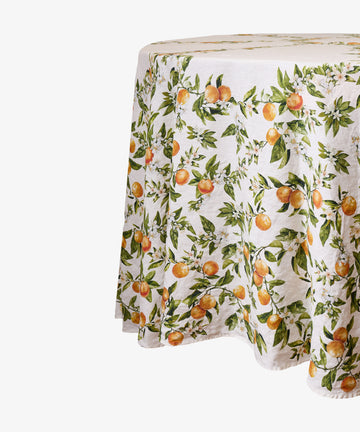 The L’Orangerie Round Linen Tablecloth covers the table with a white linen fabric adorned with a vibrant pattern of oranges and green leaves on branches, evoking classic French garden-inspired designs.
