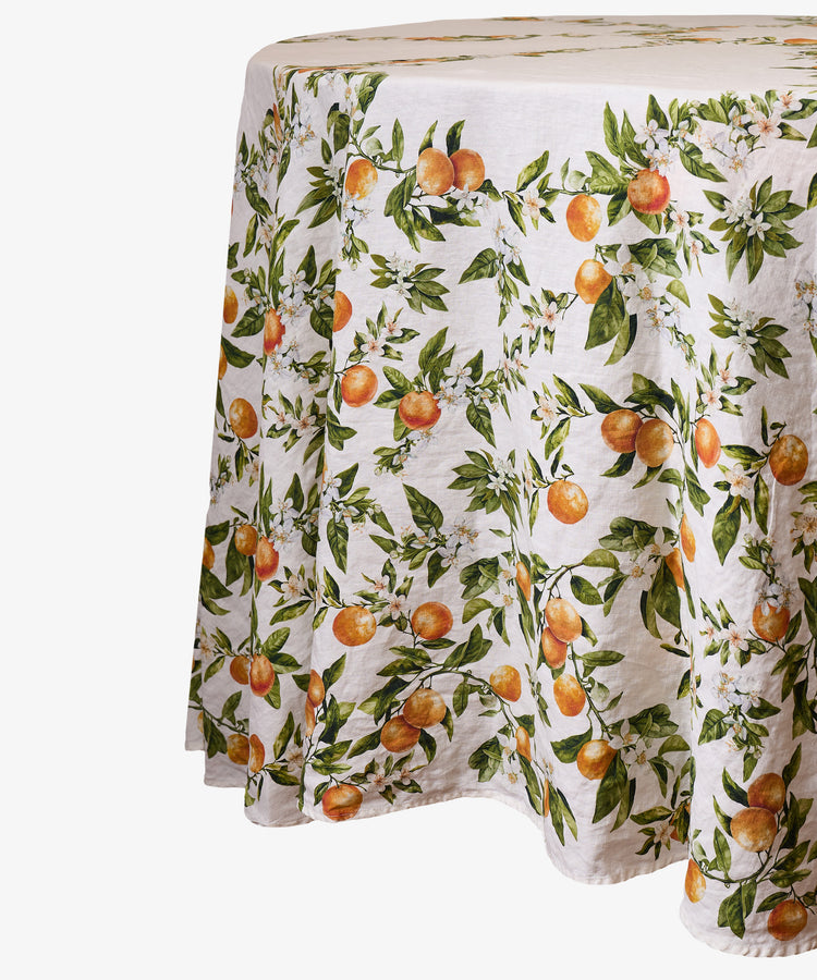 The L’Orangerie Round Linen Tablecloth drapes gracefully over the table, its white linen adorned with an orange fruit and green leaf pattern that evokes the charm of a classical French garden.