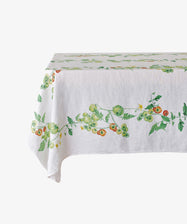 The Tomate Verte Linen Tablecloth features a botanical print of green and red tomatoes with leafy vines, designed by Summerill & Bishop, and is draped over a rectangular table.