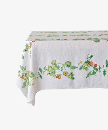 The Tomate Verte Linen Tablecloth features a botanical print of green and red tomatoes with leafy vines, designed by Summerill & Bishop, and is draped over a rectangular table.