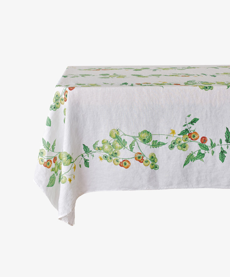 The Tomate Verte Linen Tablecloth features a botanical print of green and red tomatoes with leafy vines, designed by Summerill & Bishop, and is draped over a rectangular table.