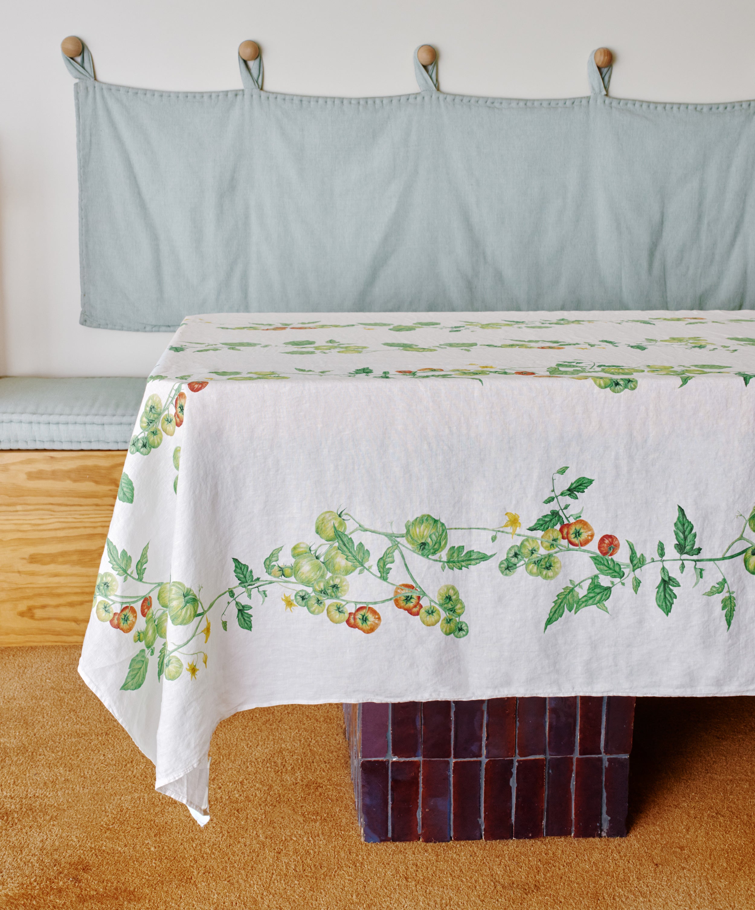 A table is dressed with the Tomate Verte Linen Tablecloth—its white linen features a tomato vine pattern—adding a beautiful Summerill & Bishop touch to a room with a light blue bench and cushion.