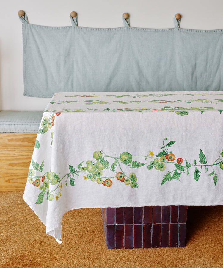 A table is dressed with the Tomate Verte Linen Tablecloth—its white linen features a tomato vine pattern—adding a beautiful Summerill & Bishop touch to a room with a light blue bench and cushion.