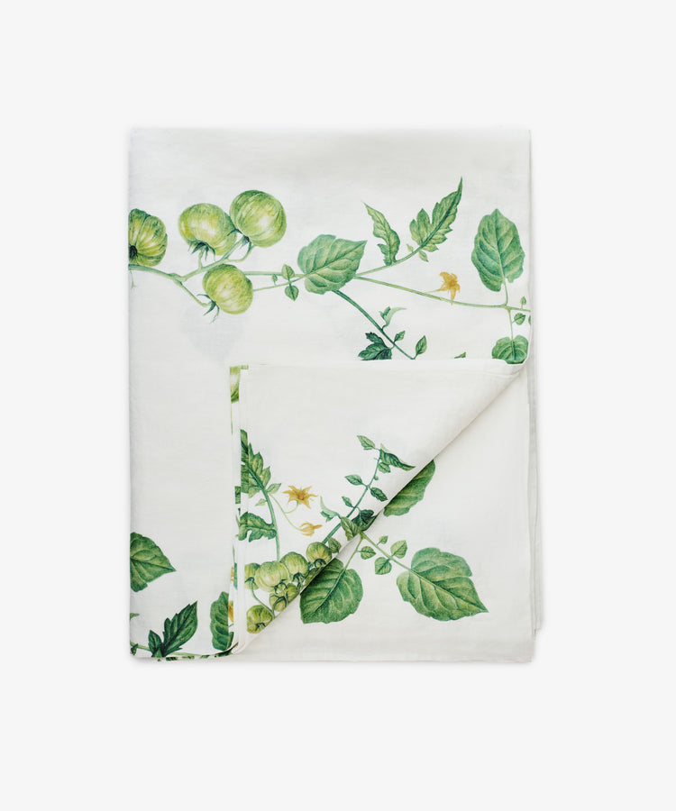 The Tomate Verte Linen Tablecloth from Summerill & Bishop features a folded white linen design with a green vine and tomato botanical print on one side.